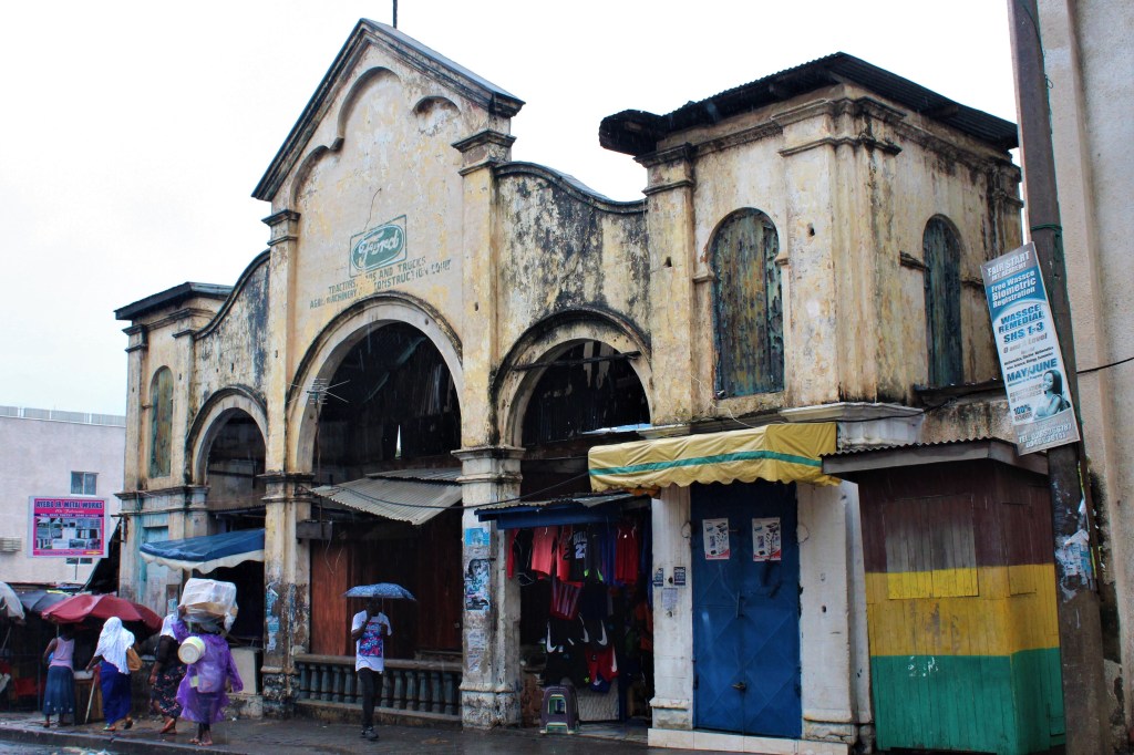 The old Ford building in old Accra on the outskirts of Makola Market, with peeling paint and an architectural style that dates back many years. It is rainy and people walk by with umbrellas. 