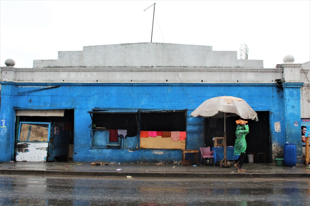 A blue building in old Accra with colourful clothes drying over the walls. It is raining and a person walks past the building in a rain coat. 
