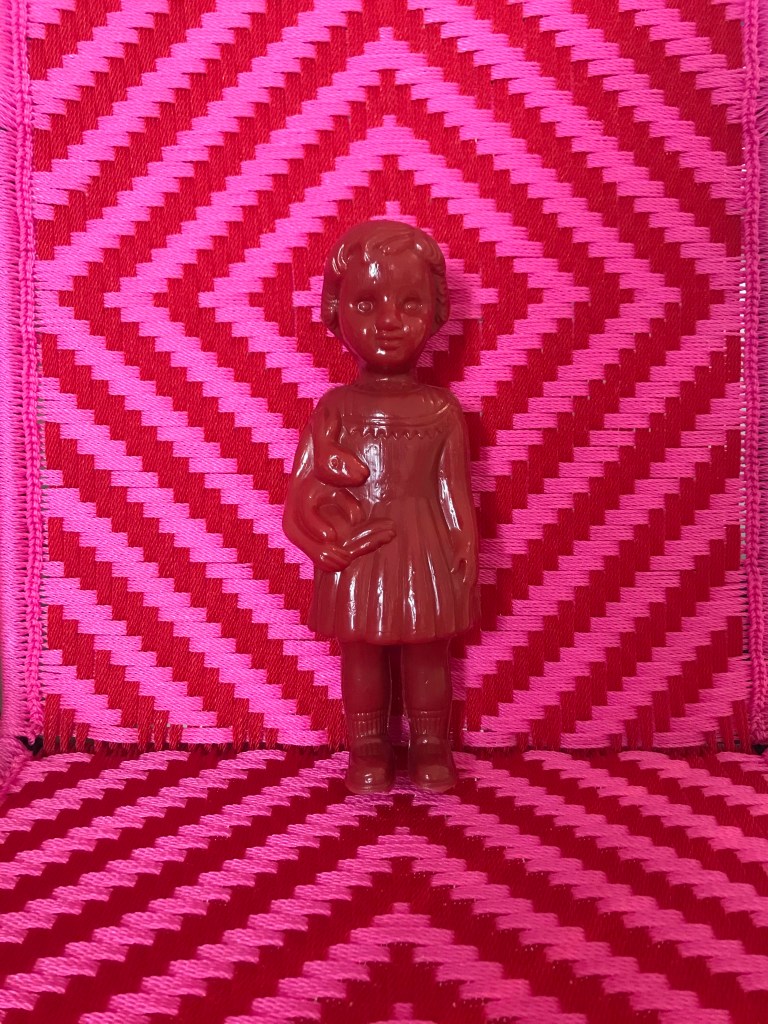 A Red Clonette Doll stands against the backdrop of a woven chair made by Joel at Calao Creation in Ghana